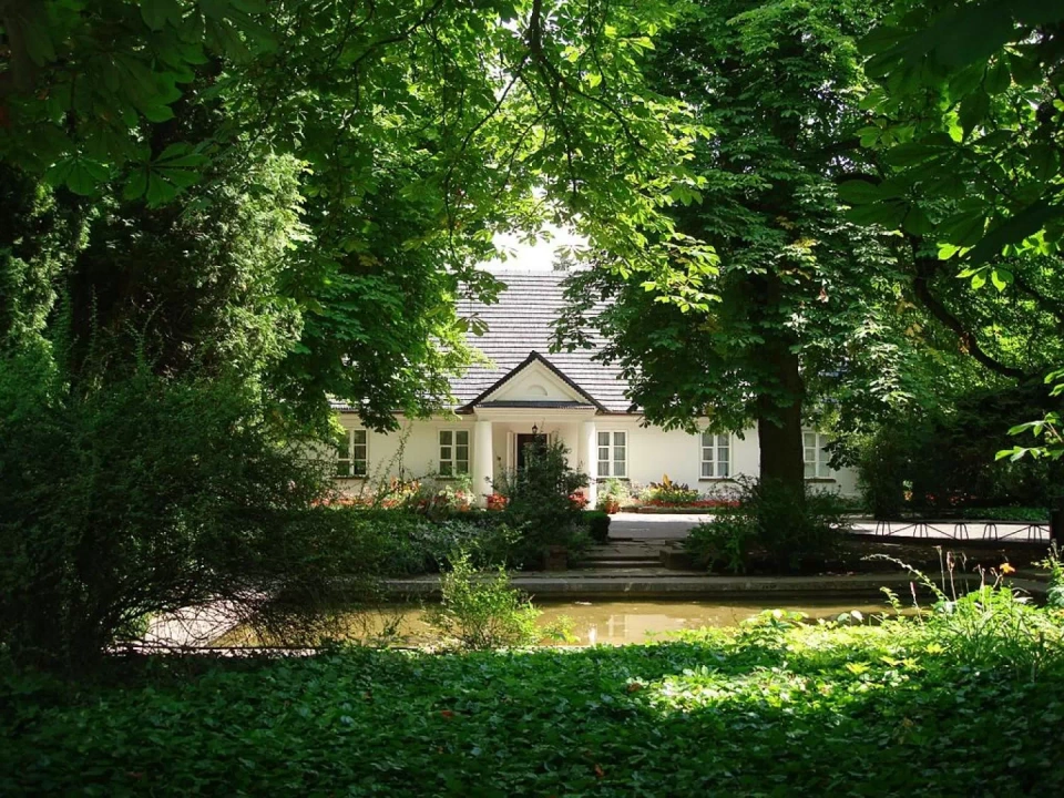 Budynek dworku w Żelazowej Woli. Minimalistyczny budynek z białą elewacją, kolumnadą przed wejściem i czarnym dachem. Przed nim znajduje się park pełen drzew i krzew&oacute;w, a także staw.